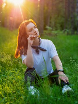 A young woman sits on the green grass in the summer in the forest against the backdrop of the setting sun. photo