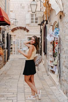 Girl tourist walking through ancient narrow street on a beautiful summer day in MEDITERRANEAN MEDIEVAL CITY , OLD TOWN bUDVA, MONTENEGRO. Young beautiful cheerful woman walking on old street at tropical town. Pretty girl looking at you and smiling