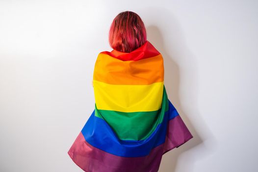 Young red-haired woman stands with her back against a white background and holds the lgbt flag.