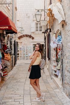 Girl tourist walking through ancient narrow street on a beautiful summer day in MEDITERRANEAN MEDIEVAL CITY , OLD TOWN bUDVA, MONTENEGRO. Young beautiful cheerful woman walking on old street at tropical town. Pretty girl looking at you and smiling