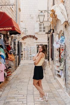 Girl tourist walking through ancient narrow street on a beautiful summer day in MEDITERRANEAN MEDIEVAL CITY , OLD TOWN bUDVA, MONTENEGRO. Young beautiful cheerful woman walking on old street at tropical town. Pretty girl looking at you and smiling