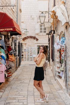 Girl tourist walking through ancient narrow street on a beautiful summer day in MEDITERRANEAN MEDIEVAL CITY , OLD TOWN bUDVA, MONTENEGRO. Young beautiful cheerful woman walking on old street at tropical town. Pretty girl looking at you and smiling