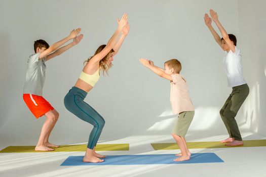 Slim woman, boy and two teenagers perform yoga exercise, stand on mats in chair pose, in sunny studio.