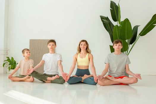 Sports family, a slender woman, a boy and two teenagers, sit in a lotus position in a bright room with green plants. copy space