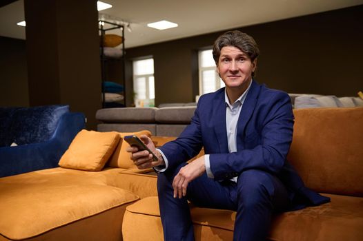 Portrait of a mature Caucasian man shopping or working in upholstered furniture warehouse, sitting on a sofa with smartphone in his hands and looking at camera