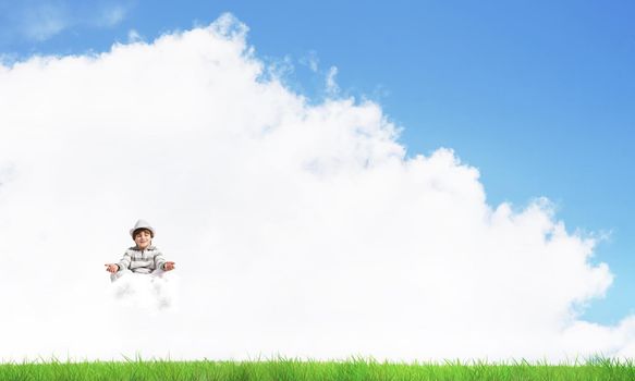Young little boy keeping eyes closed and looking concentrated while meditating on cloud in the air with bright and beautiful landscape on background.