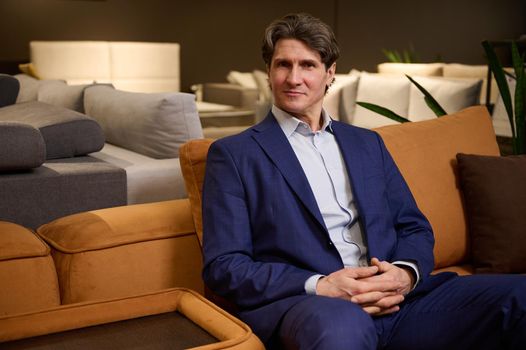 Handsome mature man in business suit, sales representative, retail assistant, interior designer, furniture store manager, business owner sitting on a comfortable sofa and confidently looking at camera