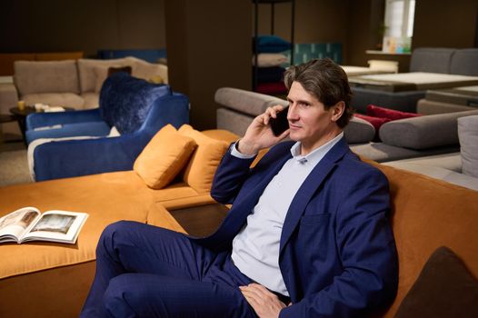 Handsome elegant imposing mature Caucasian man in a business suit talking on the phone while sitting on a sofa in a furniture store exhibition center. Home improvement and interior design concept