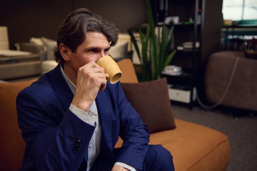 Mature man in business suit drinking coffee, sitting on a sofa in the furniture store showroom. Interior design, house remodelling, home improvement, creative occupation and small business concept