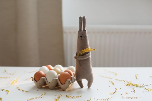 Funny handmade bunny rabbit toy with colorful chicken eggs and yellow mimosa flowers. Easter gift concept.