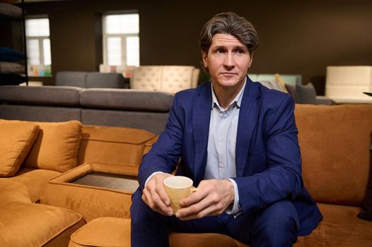 A furniture showroom client, handsome European man in business suit holding a cup of coffee, sitting on sofa and waiting for a consultation of a home decorator about the new interior design project