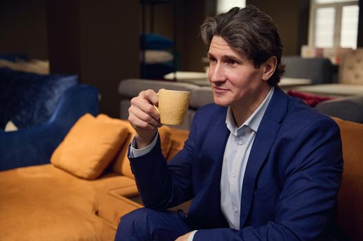 Furniture store client, handsome man in business suit enjoying coffee break sitting on the sofa and waiting for a consultation of an interior designer about the new project of home remodelling