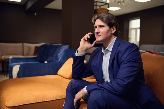 Portrait of a handsome mature European man in business suit, client customer buyer talking on mobile phone while visiting a furniture store and testing sofa. Sales manager, interior designer at work