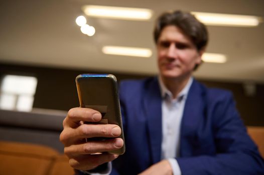 Close-up of the hands of a blurred mature businessman, sales manager holding a mobile phone, typing text messages, surfing on websites, browsing, swiping. Business, internet, communication concept