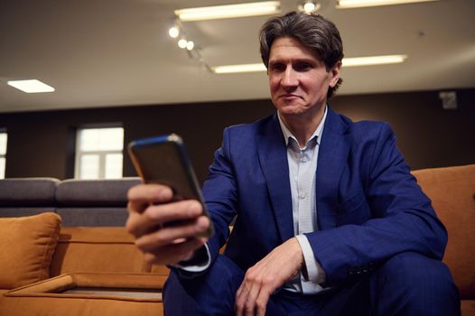 Portrait of a mature European man shopping or working in upholstered furniture warehouse, sitting on a sofa with smartphone in his hands and looking at camera