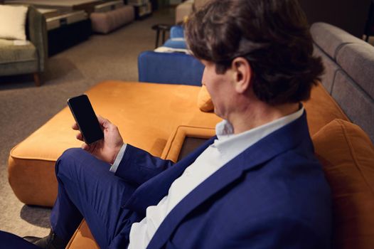 Focus on a smartphone with blank black screen with copy ad space in the hands of a mature Caucasian man in business suit, sitting on a sofa in furniture store showroom.