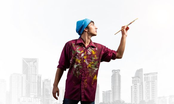 Young artist posing with paintbrush. Painter in dirty shirt and bandana standing on background modern downtown. Creative hobby and artistic occupation concept with copy space. Art classes advertising