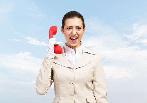 Expressive woman shouting into red handset phone. Operator in business suit standing with cellphone on blue sky background. Business assistance and consultation. Customer service support concept