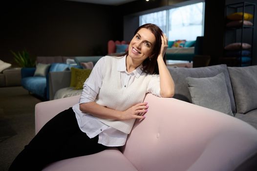 Business portrait of a charming middle aged confident Caucasian woman - interior designer, business owner sitting on stylish pink sofa in furniture store and smiling beautiful smile looking at camera