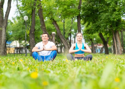Content peaceful young couple with closed eyes sitting in lotus position. Man and woman doing yoga in park together. Meditation in yoga class. Morning exercising outdoors and healthy lifestyle.