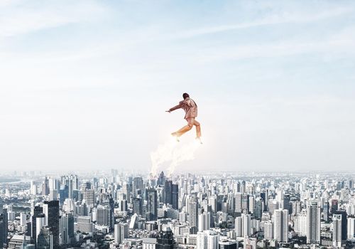 Expressive businessman in suit and aviator hat flying in blue sky as superhero. Business person as superman with jet flame above modern downtown. New challenges and winning. Career ambition and growth