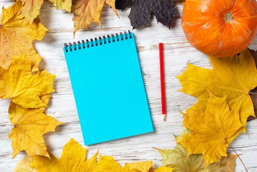 Spiral notepad with pencil and ripe pumpkin lies on vintage wooden desk with bright foliage. Happy thanksgiving holiday congratulation. Flat lay composition with autumn leaves on wooden surface.