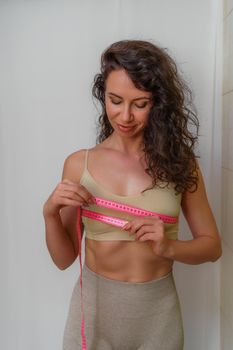 Cropped view of slim woman measuring breasts with tape measure at home, close up. Unrecognizable European woman checks the result of a weight loss diet or liposuction indoors. Healthy lifestyle..