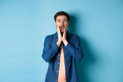 Portrait of guy gasping and dropping jaw with shocked face, staring at camera startled, standing on blue background.