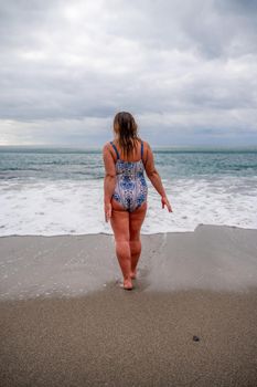 A plump woman in a bathing suit enters the water during the surf. Alone on the beach, Gray sky in the clouds, swimming in winter