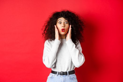 Surprised young woman hear amazing news, looking at promo and saying wow, stare with disbelief, standing on red background.