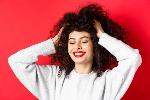 Close-up of carefree woman touching soft curly hair and smiling pleased, standing on red background. Haircare and beauty concept.