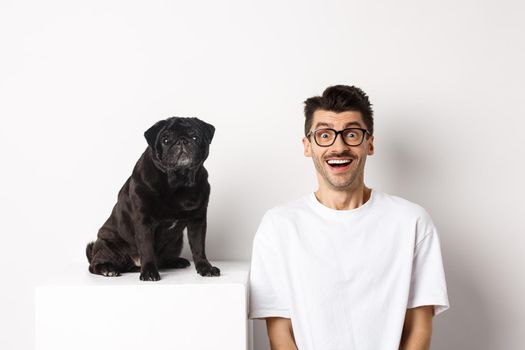 Cheerful young man in glasses sitting near cute small dog and looking at camera. Dog owner staring happy and amazed, white background.