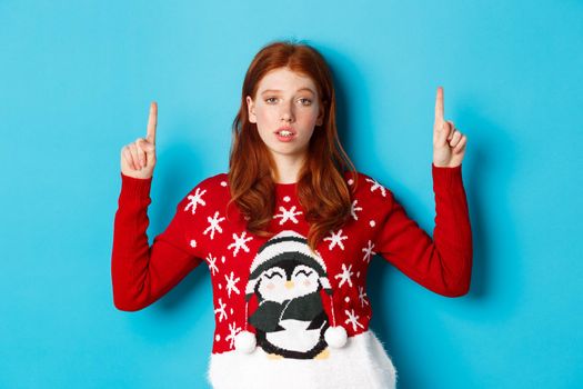 Merry Christmas. Skeptical and unamused redhead girl pointing fingers up, showing logo with reluctant face, standing over blue background.