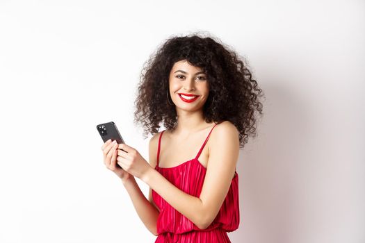 Happy elegant woman in red dress writing message, using smartphone and smiling at camera, standing against white background.