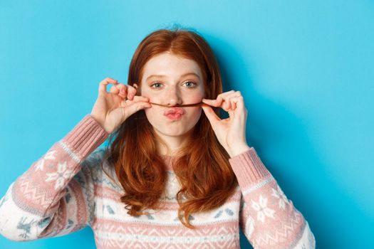 Close-up of silly and funny redhead girl making moustache with hair strand and puckered lips, grimacing against blue background.