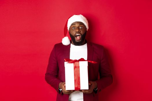Christmas, New Year and shopping concept. Excited Black man receiving xmas gift, looking surprised, holding wrapped present and staring at camera, wearing santa hat.