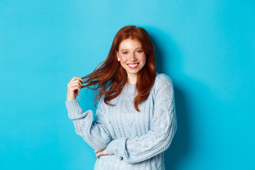 Flirty young woman with red hair, playing with hair and smiling, standing in sweater against blue background.
