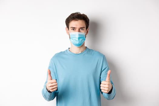 Covid-19, pandemic and social distancing concept. Young man in casual clothes and medical mask, showing thumbs up and looking serious, protecting from virus during quarantine.