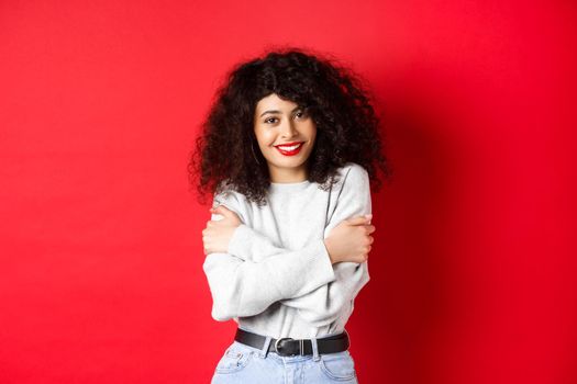 Tender young woman hugging herself, feeling comfortable and happy, smiling silly at camera, standing against red background.