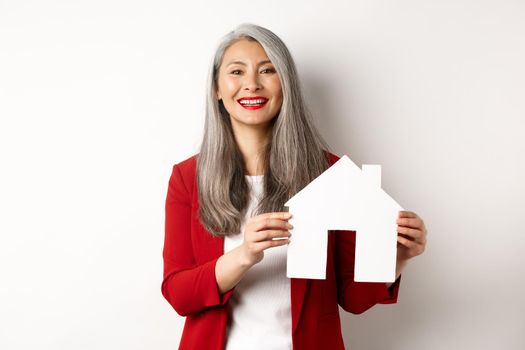 Asian female real estate agent showing paper house cutout, broker smiling friendly and selling property, standing over white background.