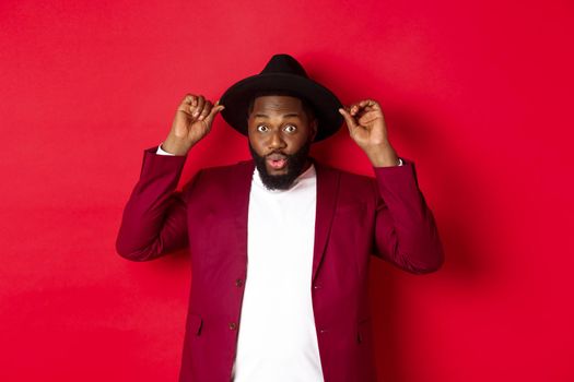 Fashion and party concept. Handsome Black man wearing hat going on holiday celebration, looking excited, standing over red background.