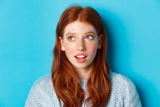 Headshot of thoughtful redhead teen girl looking at upper left corner, staring at logo with curious expression, standing over blue background.