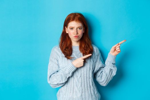 Winter holidays and people concept. Complicated redhead girl staring indecisive, pointing fingers right at logo and frowning, standing perplexed against blue background.