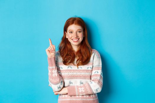 Excited redhead teen girl having an idea, raising finger and smiling, suggesting something, standing in sweater over blue background.