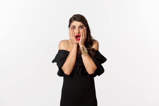 Fashion and beauty. Shocked and worried woman in black dress looking scared, standing anxious over white background.