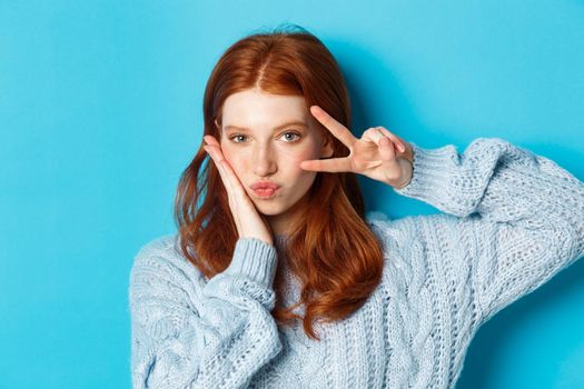 Close-up of beautiful and sassy girl with red hair, showing peace kawaii sign and gazing at camera, standing over blue background.
