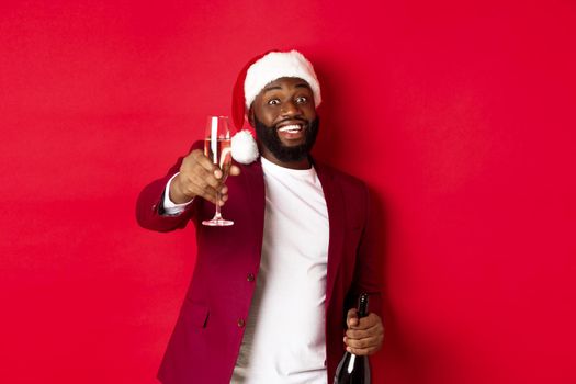 Christmas, party and holidays concept. Handsome Black man in santa hat raising glass of champagne and smiling, saying toast, celebrating New Year, red background.