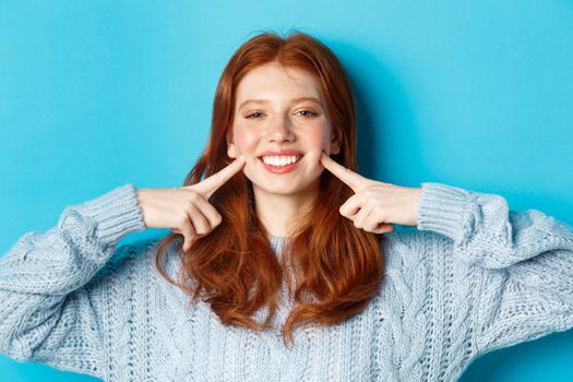 Close-up of cheerful teenage girl with red hair and freckles, poking cheeks, showing dimples and smiling with white teeth, standing over blue background.