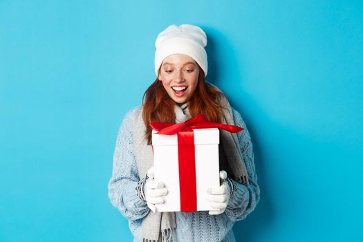 Winter holidays and Christmas eve concept. Surprised cute redhead girl in beanie and sweater receiving New Year gift, looking at present amazed, standing over blue background.