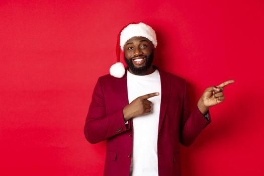 Christmas, party and holidays concept. Smiling Black man with beard and santa hat, pointing fingers right at showing logo, standing over red background.